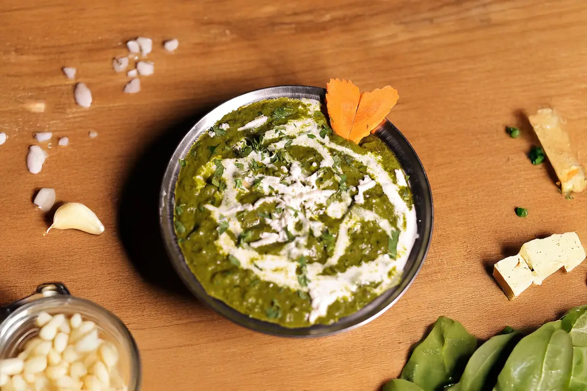 Palak Paneer in einer Stahlschale auf warmem Eichenholz, daneben Paneerwürfel und ein Sahnespiegel.